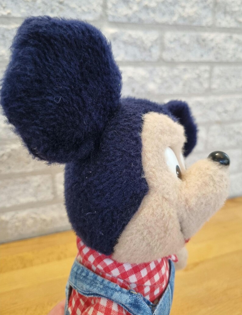 May include: A plush Mickey Mouse toy with navy blue ears, a tan face, and a black nose. He wears a red and white checkered shirt and denim overalls. The toy is soft and cuddly.