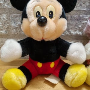 May include: A plush Mickey Mouse toy with black ears, a black nose, and yellow shoes. The toy is wearing red shorts with two yellow buttons.