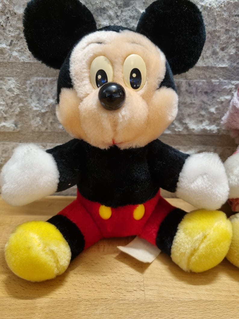 May include: A plush Mickey Mouse toy with black ears, a black nose, and yellow shoes. The toy is wearing red shorts with two yellow buttons.