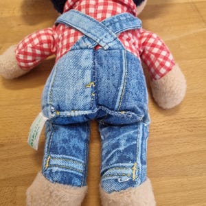 May include: A stuffed doll with a dark blue head, red and white checkered shirt, and blue denim overalls. The doll has tan-colored hands and feet. The overalls have yellow stitching and the doll is viewed from the back.