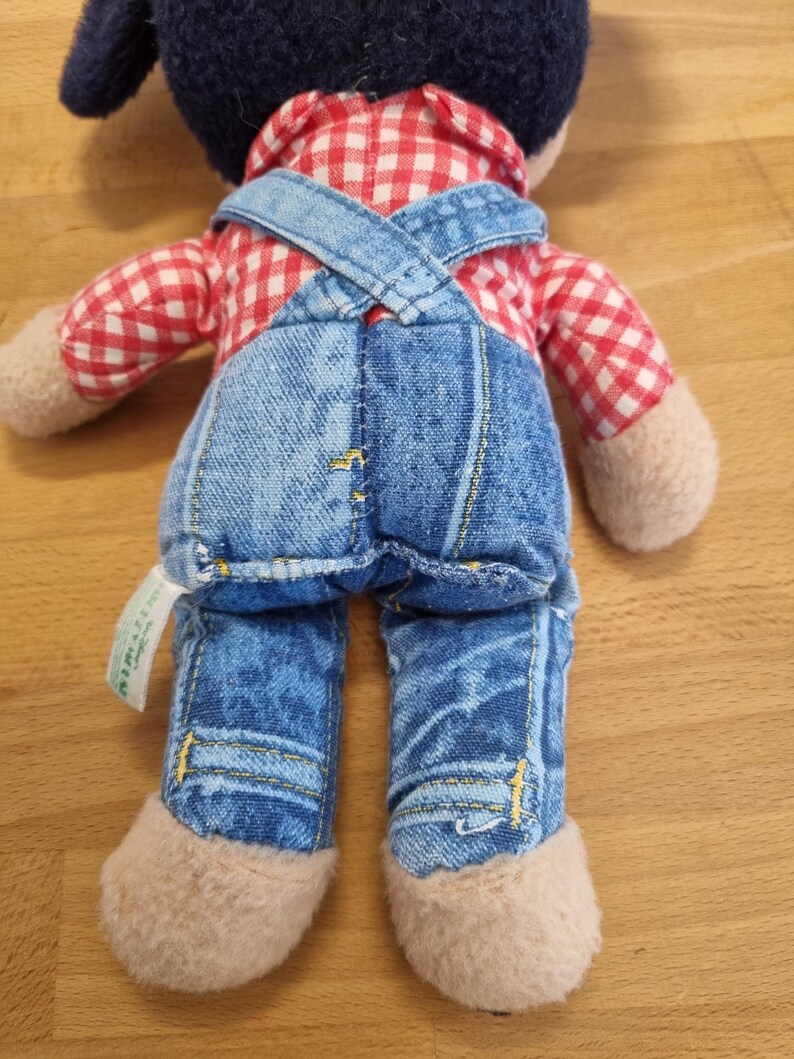 May include: A stuffed doll with a dark blue head, red and white checkered shirt, and blue denim overalls. The doll has tan-colored hands and feet. The overalls have yellow stitching and the doll is viewed from the back.