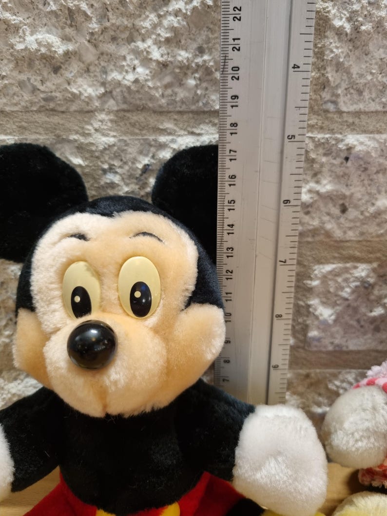 May include: A plush Mickey Mouse toy with black ears, a black nose, and large yellow eyes. The toy is wearing red shorts with yellow buttons. The toy is approximately 11 inches tall.