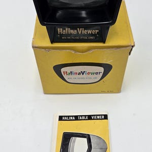 May include: A vintage Halina Viewer, with a black and white design, sits atop its original yellow box. The box and viewer feature the "Halina Viewer" logo and text. A booklet with the same logo is also present.