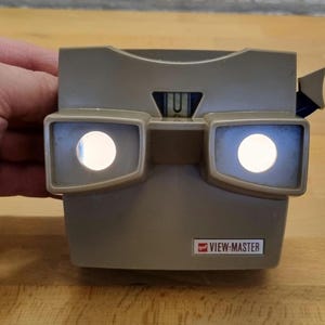 Könnte beinhalten: Ein beigefarbener View-Master-Betrachter mit zwei beleuchteten Okularen. Das klassische Spielzeug hat eine rechteckige Form mit einem kleinen rot-weißen View-Master-Logo auf der Vorderseite. Der Betrachter liegt auf einer Holzoberfläche.