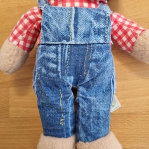 May include: A plush doll wearing blue denim overalls and a red and white checkered shirt. The doll has beige colored hands and feet. The overalls have yellow stitching. The doll is on a light brown surface.