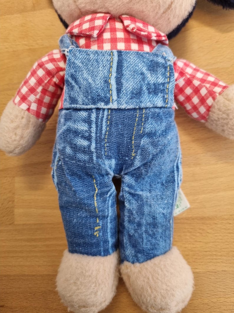 May include: A plush doll wearing blue denim overalls and a red and white checkered shirt. The doll has beige colored hands and feet. The overalls have yellow stitching. The doll is on a light brown surface.