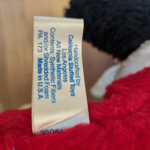 May include: A white tag with blue text attached to a red stuffed toy. The tag reads "Handcrafted by California Stuffed Toys Los Angeles All New Materials Contents: Synthetic Fibers and/or Shredded Foam PA. 173 Made in U.S.A. 30064"