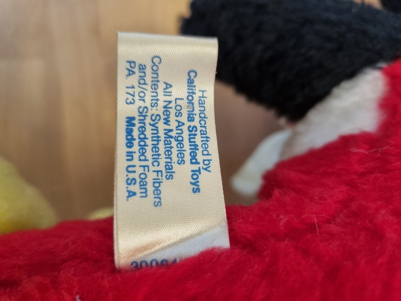 May include: A white tag with blue text attached to a red stuffed toy. The tag reads "Handcrafted by California Stuffed Toys Los Angeles All New Materials Contents: Synthetic Fibers and/or Shredded Foam PA. 173 Made in U.S.A. 30064"