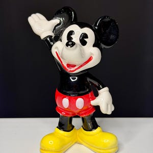K&ouml;nnte beinhalten: Eine Keramikfigur von Mickey Mouse. Die Figur ist schwarz, wei&szlig;, rot und gelb. Mickey winkt mit seiner rechten Hand. Er tr&auml;gt rote Shorts mit wei&szlig;en Kn&ouml;pfen und gelbe Schuhe. Die Figur hat ein gl&auml;nzendes Finish.