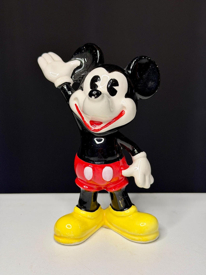 K&ouml;nnte beinhalten: Eine Keramikfigur von Mickey Mouse. Die Figur ist schwarz, wei&szlig;, rot und gelb. Mickey winkt mit seiner rechten Hand. Er tr&auml;gt rote Shorts mit wei&szlig;en Kn&ouml;pfen und gelbe Schuhe. Die Figur hat ein gl&auml;nzendes Finish.