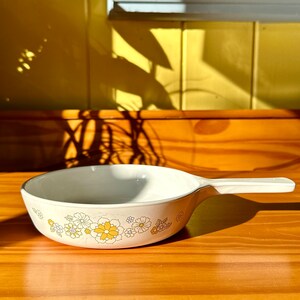 May include: A white ceramic casserole dish with a floral pattern in yellow and blue. The dish has a single handle and is sitting on a wooden surface.