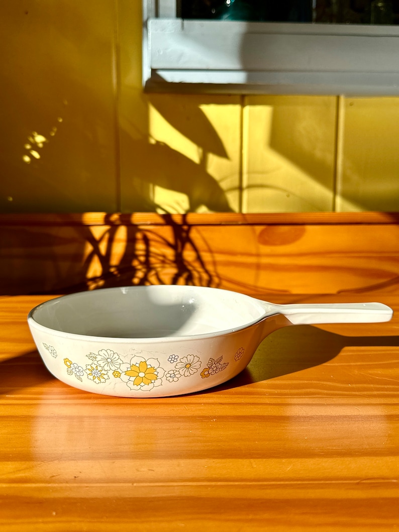May include: A white ceramic casserole dish with a floral pattern in yellow and blue. The dish has a single handle and is sitting on a wooden surface.