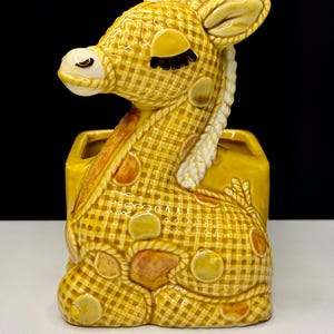 May include: A ceramic giraffe planter in shades of yellow and brown. The giraffe has a checkered pattern on its body and is in a seated position. The planter has a rectangular base and a white braided mane.