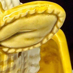 May include: Close-up of a yellow ceramic figurine, possibly a vintage piece. The figurine has a detailed, textured surface with a ruffled edge and a white accent. The yellow color is consistent throughout the piece, with some areas showing wear.