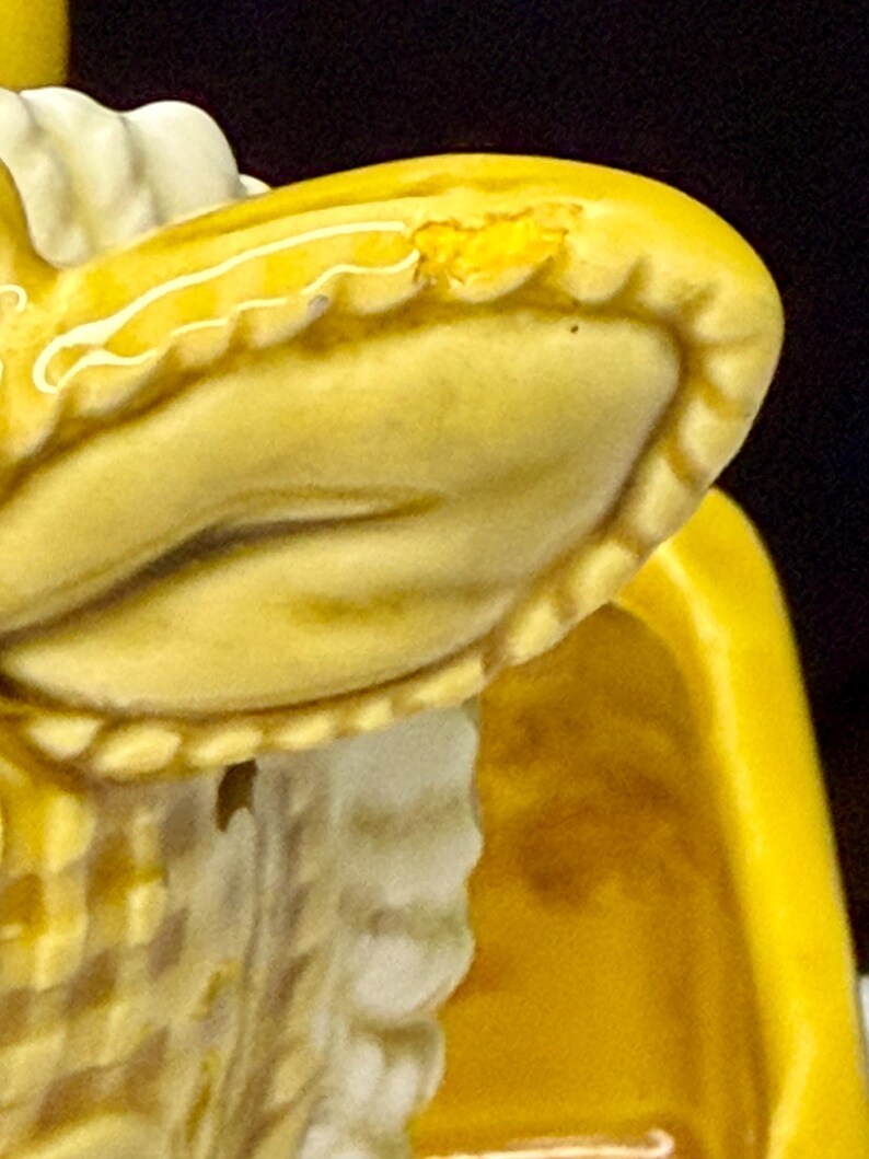 May include: Close-up of a yellow ceramic figurine, possibly a vintage piece. The figurine has a detailed, textured surface with a ruffled edge and a white accent. The yellow color is consistent throughout the piece, with some areas showing wear.