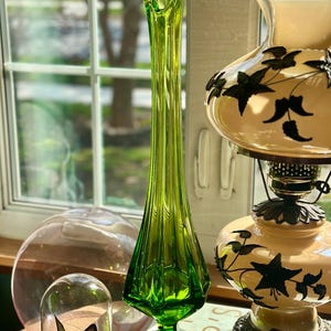 May include: A tall, green glass vase with a flared opening and a faceted design. The vase is sitting on a table with a book and a lamp.