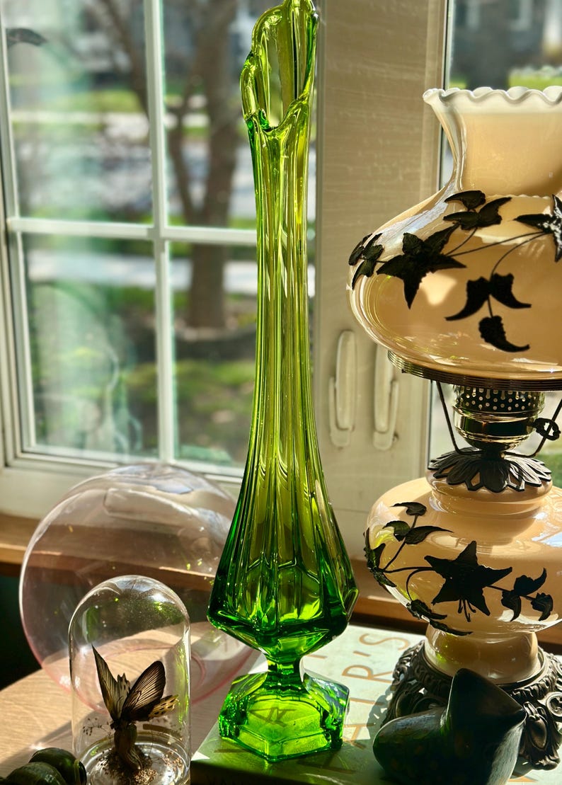 May include: A tall, green glass vase with a flared opening and a faceted design. The vase is sitting on a table with a book and a lamp.