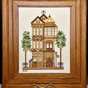 May include: A framed cross-stitch embroidery depicting a colorful Victorian-style house with a decorative roof and two trees. The embroidery is signed "L.a. 2000" in the bottom left corner.