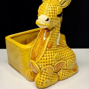 May include: A yellow ceramic giraffe planter with a checkered pattern. The giraffe is lying down with its head up, and the planter has a rectangular shape. The giraffe has brown accents and white details.