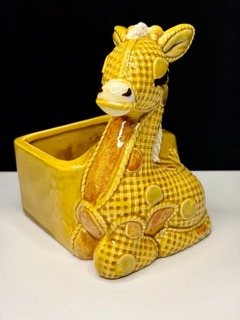 May include: A yellow ceramic giraffe planter with a checkered pattern. The giraffe is lying down with its head up, and the planter has a rectangular shape. The giraffe has brown accents and white details.