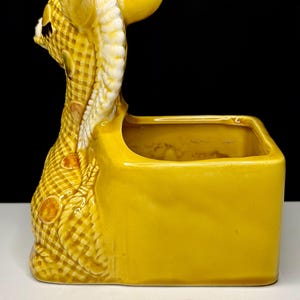 May include: A yellow ceramic planter in the shape of a giraffe. The giraffe's head and neck are detailed with a checkered pattern and white accents. The planter has a rectangular shape.