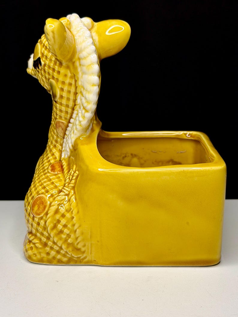 May include: A yellow ceramic planter in the shape of a giraffe. The giraffe's head and neck are detailed with a checkered pattern and white accents. The planter has a rectangular shape.