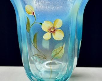 May include: A light blue glass vase with a hand-painted floral design. The vase has a unique, wavy shape and features yellow flowers and green leaves.