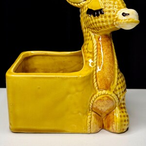 May include: A yellow ceramic donkey planter with a rectangular container and a decorative rope design. The donkey has a checkered pattern, closed eyes, and a white muzzle. The planter is a decorative item.