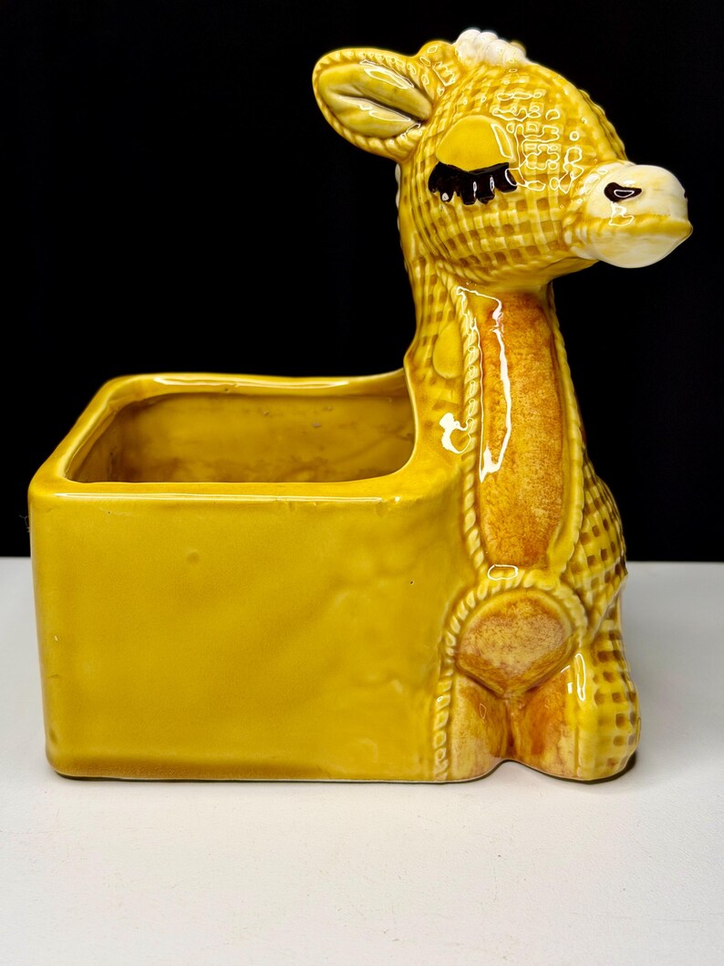 May include: A yellow ceramic donkey planter with a rectangular container and a decorative rope design. The donkey has a checkered pattern, closed eyes, and a white muzzle. The planter is a decorative item.
