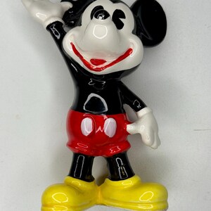 K&ouml;nnte beinhalten: Eine Keramikfigur von Mickey Mouse, einer klassischen Disney-Figur. Mickey wird mit einem schwarzen K&ouml;rper, roten Shorts, gelben Schuhen und wei&szlig;en Handschuhen dargestellt. Sein rechter Arm ist erhoben, und er hat einen fr&ouml;hlichen Gesichtsausdruck.