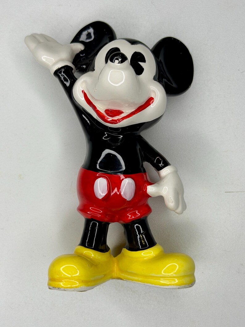 K&ouml;nnte beinhalten: Eine Keramikfigur von Mickey Mouse, einer klassischen Disney-Figur. Mickey wird mit einem schwarzen K&ouml;rper, roten Shorts, gelben Schuhen und wei&szlig;en Handschuhen dargestellt. Sein rechter Arm ist erhoben, und er hat einen fr&ouml;hlichen Gesichtsausdruck.