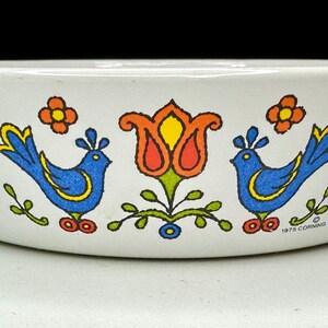 May include: A white ceramic baking dish with a blue and yellow bird design, a red and yellow tulip, and green leaves. The dish has the text "1975 CORNING" on the bottom.