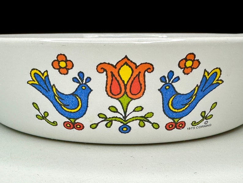 May include: A white ceramic baking dish with a blue and yellow bird design, a red and yellow tulip, and green leaves. The dish has the text "1975 CORNING" on the bottom.