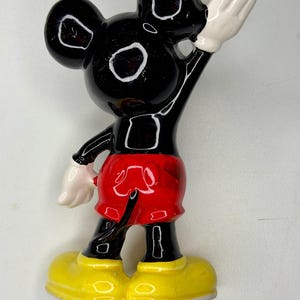 K&ouml;nnte beinhalten: Eine schwarze Mickey Mouse-Figur mit wei&szlig;en Handschuhen und gelben Schuhen. Die Figur tr&auml;gt rote Shorts. Die Figur winkt mit der rechten Hand. Die Figur hat wei&szlig;e Augen und Ohren.