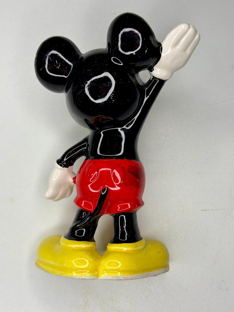K&ouml;nnte beinhalten: Eine schwarze Mickey Mouse-Figur mit wei&szlig;en Handschuhen und gelben Schuhen. Die Figur tr&auml;gt rote Shorts. Die Figur winkt mit der rechten Hand. Die Figur hat wei&szlig;e Augen und Ohren.