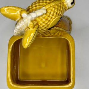 May include: A ceramic ashtray with a detailed lobster design in shades of yellow and white. The lobster is perched atop a rectangular tray, creating a unique and decorative piece. The ashtray is a functional item with a vintage aesthetic.