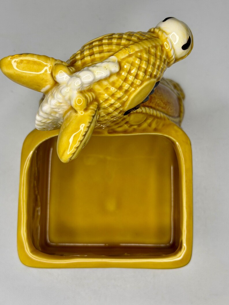 May include: A ceramic ashtray with a detailed lobster design in shades of yellow and white. The lobster is perched atop a rectangular tray, creating a unique and decorative piece. The ashtray is a functional item with a vintage aesthetic.