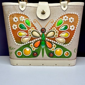 May include: A vintage beige handbag with a butterfly design. The butterfly is adorned with colorful gems, pearls, and floral accents. The bag has a cream-colored handle, clasp, and trim. The text "Butterfly" is visible on the side.
