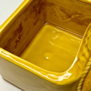 May include: A close-up of a yellow ceramic container with a rectangular shape. The interior has a glossy finish, and the exterior has a matte finish. The container has a decorative rope-like design on the side.