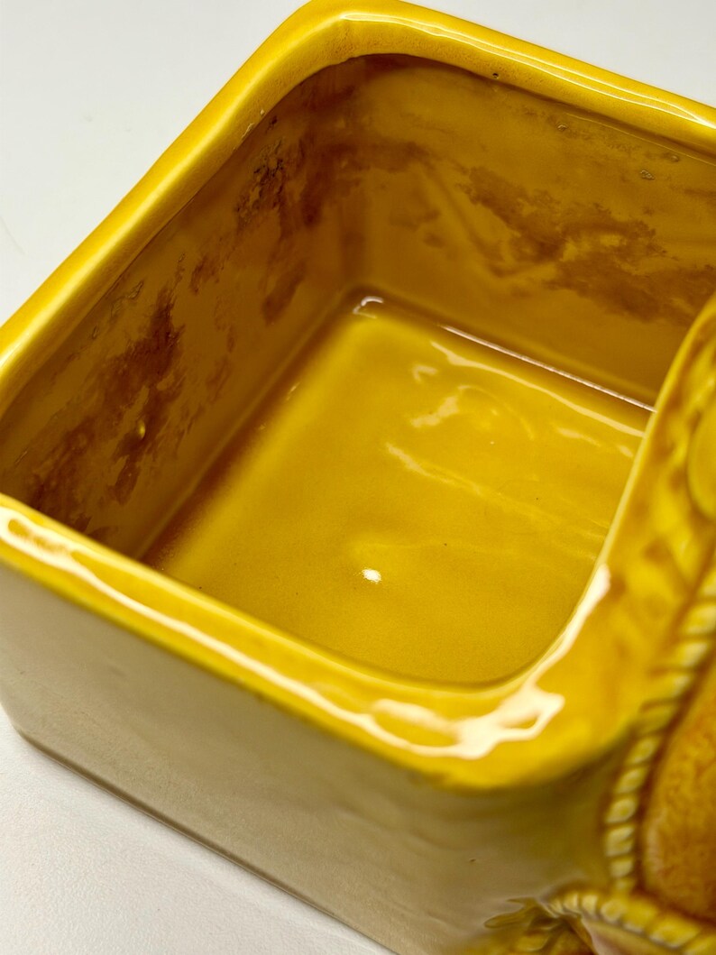 May include: A close-up of a yellow ceramic container with a rectangular shape. The interior has a glossy finish, and the exterior has a matte finish. The container has a decorative rope-like design on the side.