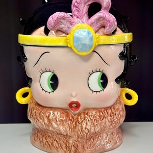 May include: A ceramic cookie jar in the shape of a cartoon character's face. The jar features a pink face, black hair, green eyes, and a pink feather-like adornment. It has a yellow headband and earrings, and a brown fur-like collar.