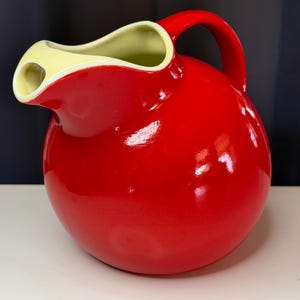 May include: A vibrant red ceramic pitcher with a rounded body and a curved handle. The interior and rim of the spout are a contrasting pale yellow. This pitcher is a decorative and functional kitchenware item.