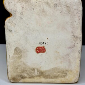 May include: A vintage ceramic figurine with a textured, off-white body and a yellow-gold trim. The base has a rectangular shape with a faded appearance. The figurine has a red and white sticker that reads "Lefton Trade Mark Exclusives Japan" and the number "H6170".