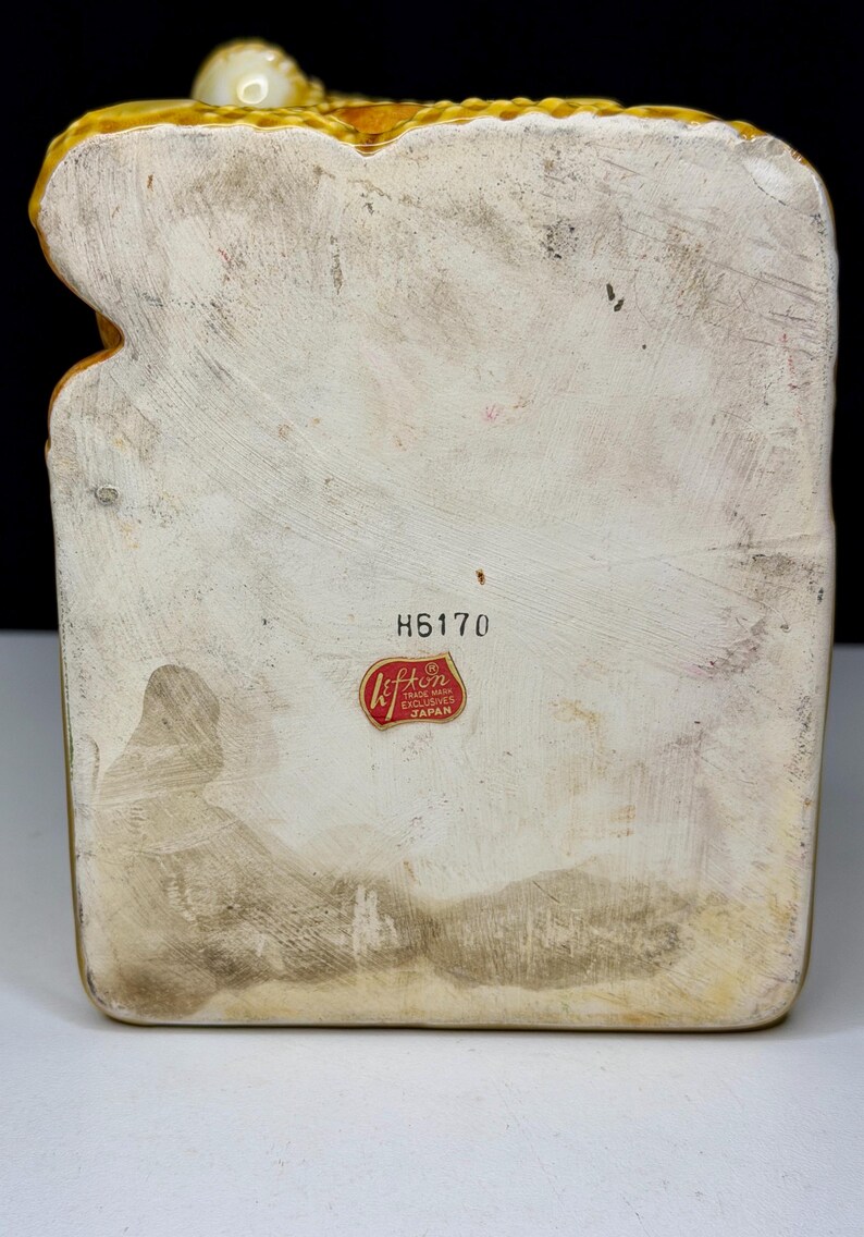 May include: A vintage ceramic figurine with a textured, off-white body and a yellow-gold trim. The base has a rectangular shape with a faded appearance. The figurine has a red and white sticker that reads "Lefton Trade Mark Exclusives Japan" and the number "H6170".