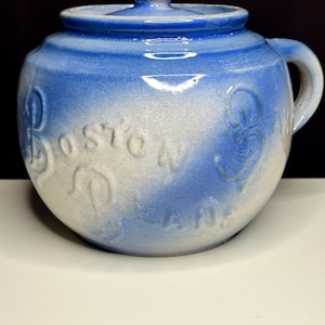 May include: A blue and white ceramic bean pot with a lid and handle. The words "Boston Beans" are embossed on the side. The pot has a rounded shape and a glossy finish, with a small knob on the lid.