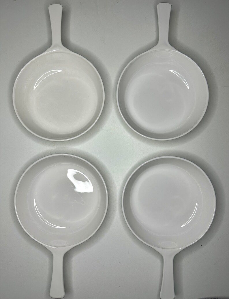 May include: Four white ceramic baking dishes with handles. The dishes are empty and arranged in a square pattern.