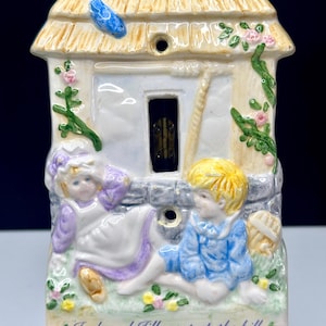 May include: A ceramic cottage figurine featuring Jack and Jill. The cottage has a thatched roof, a blue bird, and climbing flowers. Jack and Jill are depicted in blue and purple clothing, with the text "Jack and Jill went up the hill to fetch a pail of water..."