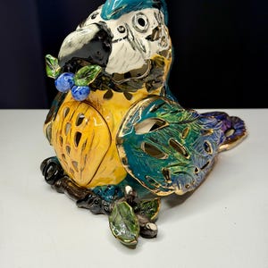 May include: A ceramic parrot figurine featuring a glossy finish with vibrant blue, yellow, and green plumage. The bird is perched on a dark brown branch with green leaves and blue berries, and includes cut-out details on its chest and wings.