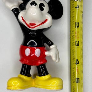 K&ouml;nnte beinhalten: Eine Keramikfigur von Mickey Mouse, mit schwarzem K&ouml;rper, roter Hose und gelben Schuhen. Die Figur ist etwa 10 cm hoch, mit wei&szlig;en Handschuhen und einem l&auml;chelnden Gesicht. Die Figur posiert mit einem erhobenen Arm.