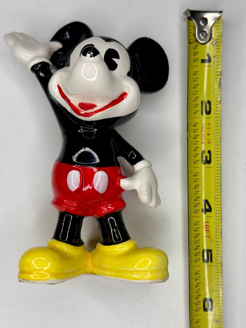 K&ouml;nnte beinhalten: Eine Keramikfigur von Mickey Mouse, mit schwarzem K&ouml;rper, roter Hose und gelben Schuhen. Die Figur ist etwa 10 cm hoch, mit wei&szlig;en Handschuhen und einem l&auml;chelnden Gesicht. Die Figur posiert mit einem erhobenen Arm.